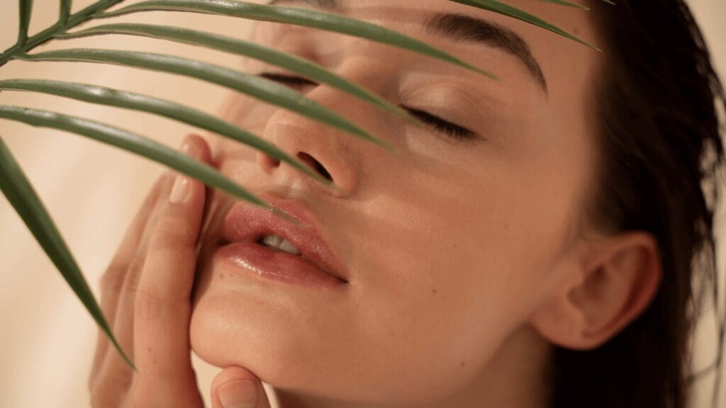 Close-up of a woman with closed eyes touching her lips, partially covered by a green leaf for a natural skincare concept.