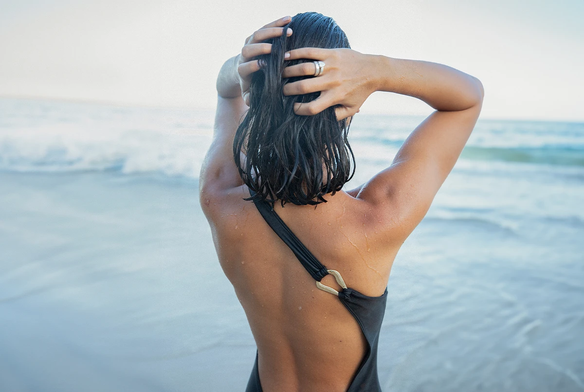 Person with wet hair looks back while standing by the ocean, wearing a black swimsuit