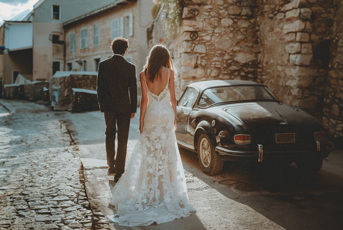 A couple walking away on a cobblestone street, with a vintage car parked beside them