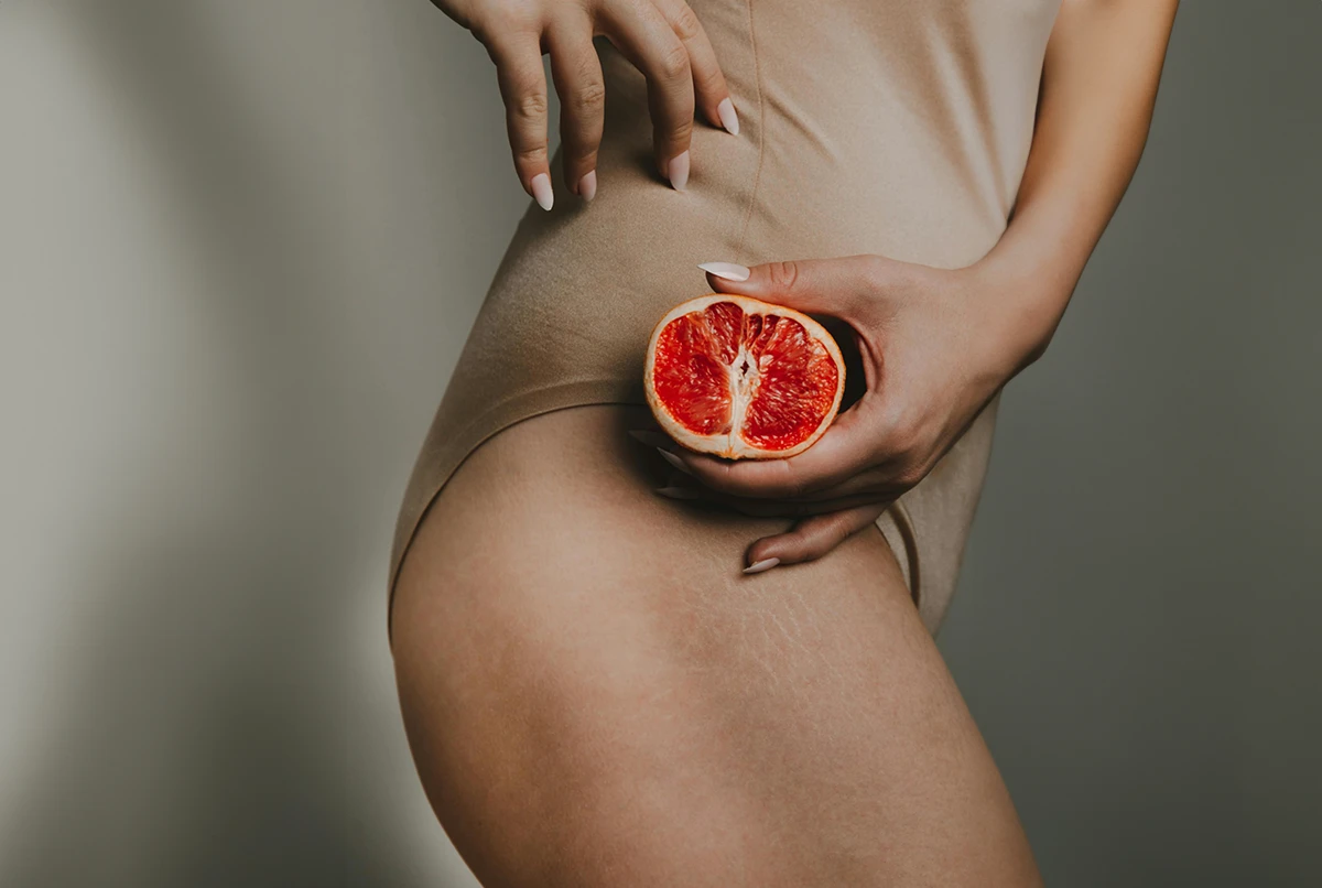 A person holds a halved red fruit near their thigh, wearing a skin-toned bodysuit