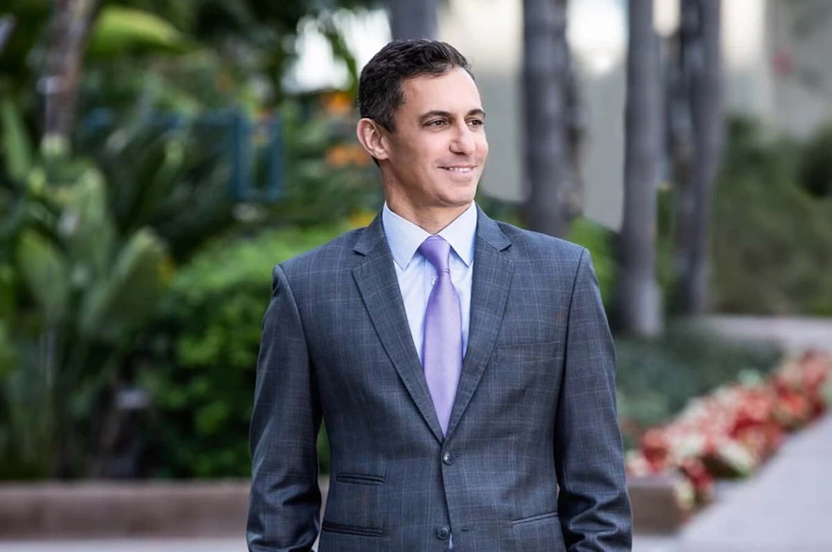 Man in a suit stands outdoors, smiling, with greenery and flowers in the background