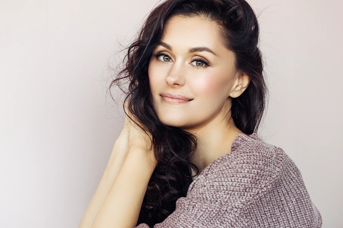 A woman with long dark hair smiles softly, resting her hand on her shoulder