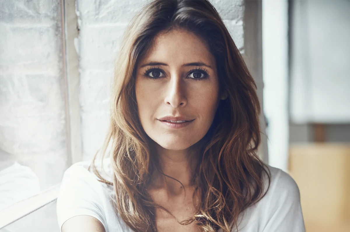 A woman with long brown hair looks directly at the camera, sitting by a window