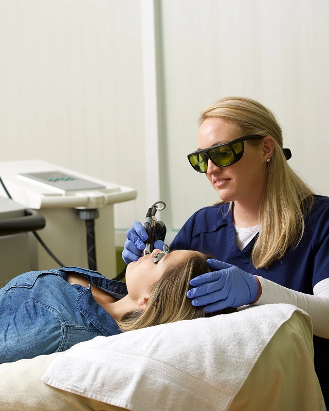 A woman in gloves uses a laser device on another woman's eyebrow while both wear protective glasses