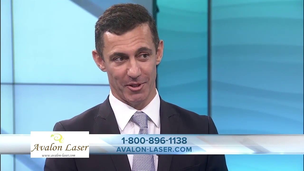 A man in a suit speaks in front of a light blue background. Contact information is displayed
