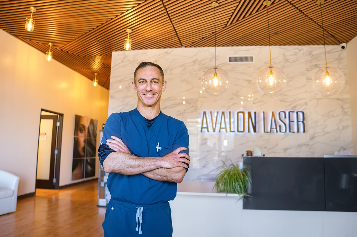 A man in scrubs stands with arms crossed in a modern clinic reception area
