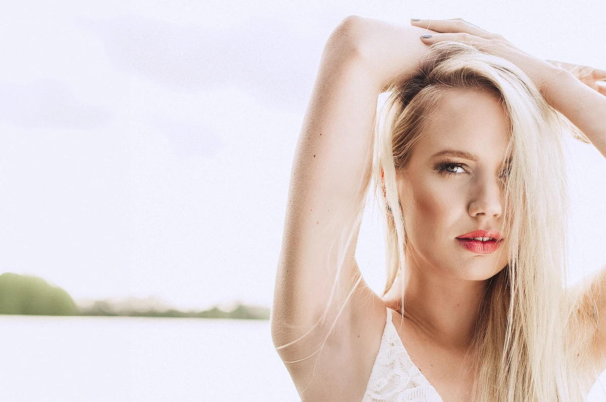 A person with long blonde hair poses with arms raised against a light background