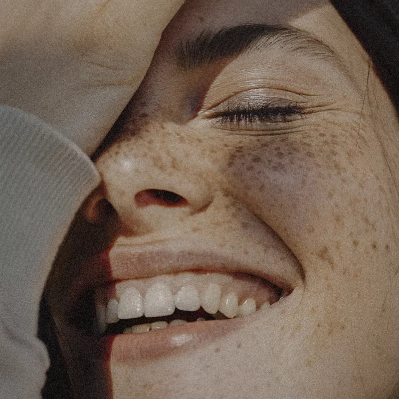 Close-up of a smiling person with freckles, eyes closed, hand covering part of the face